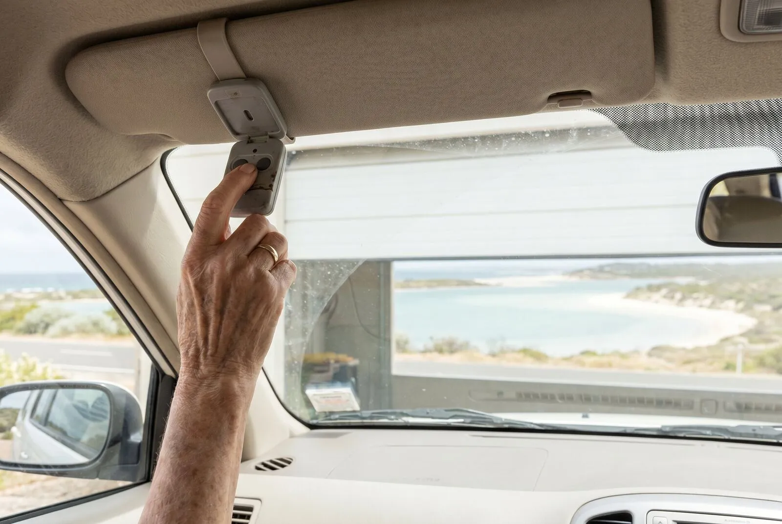 A hand pressing a garage remote on a car sun visor as the sectional door rises ahead
